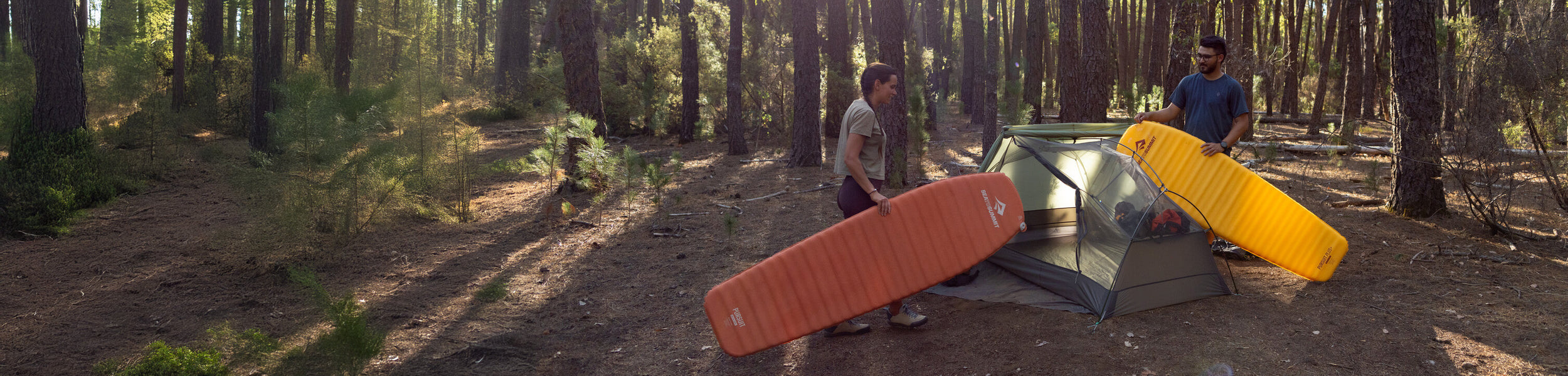 A woman and a man are holding the Pursuit and Pursuit Plus sleeping mats. They are about to put them into the Telos TR2 tent in a forest.