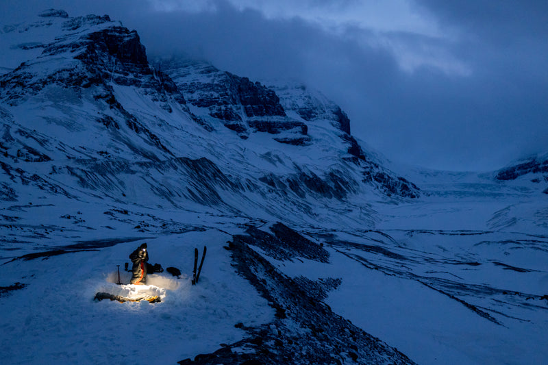  A solo backcountry skier sets up camp in a snowy, mountainous area at twilight