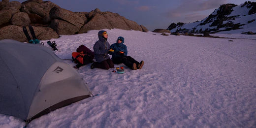 A couple of backcountry skiers laugh together at a snowy camp as they have dinner using ultralight collapsible dinnerware