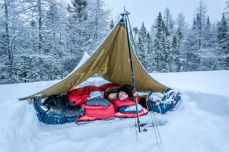 A woman lies under a tent roof in her thick sleeping bag on her sleeping mat in a winter landscape covered in snow 