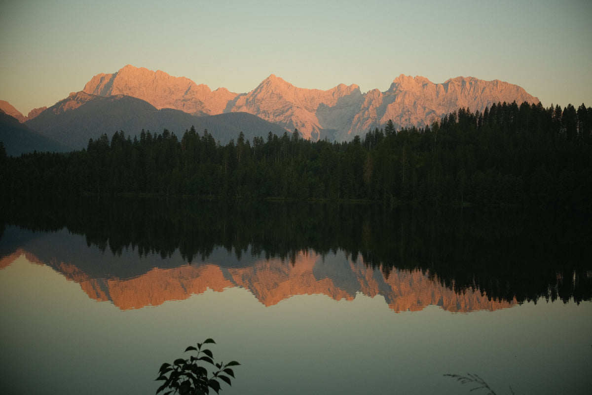 Sunlit mountain range reflected in a calm lake, surrounded by forest in the foreground.