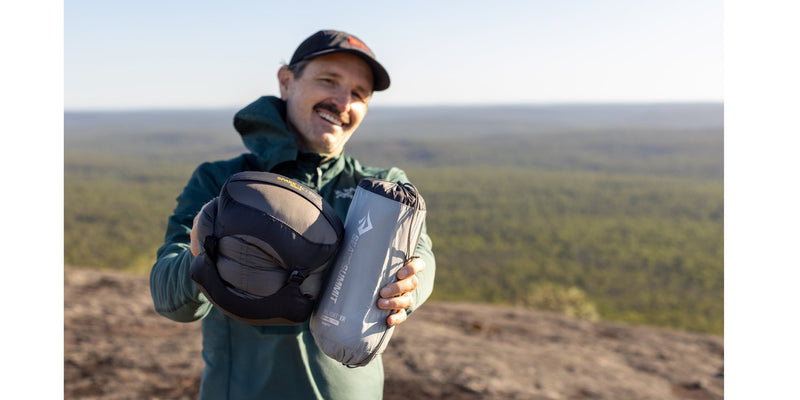 Person outdoors holding a packed Ether Light sleeping mat towards the camera