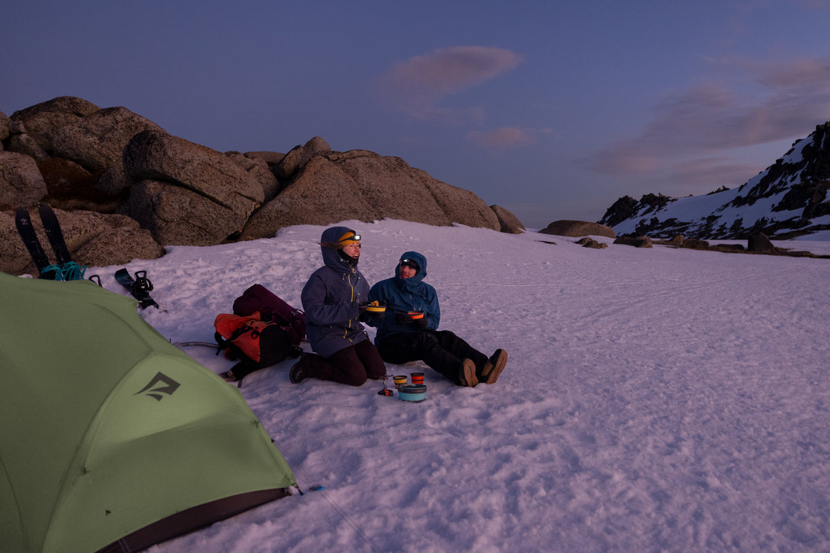 A couple of backcountry skiers laugh together at a snowy camp as they have dinner using ultralight collapsible dinnerware