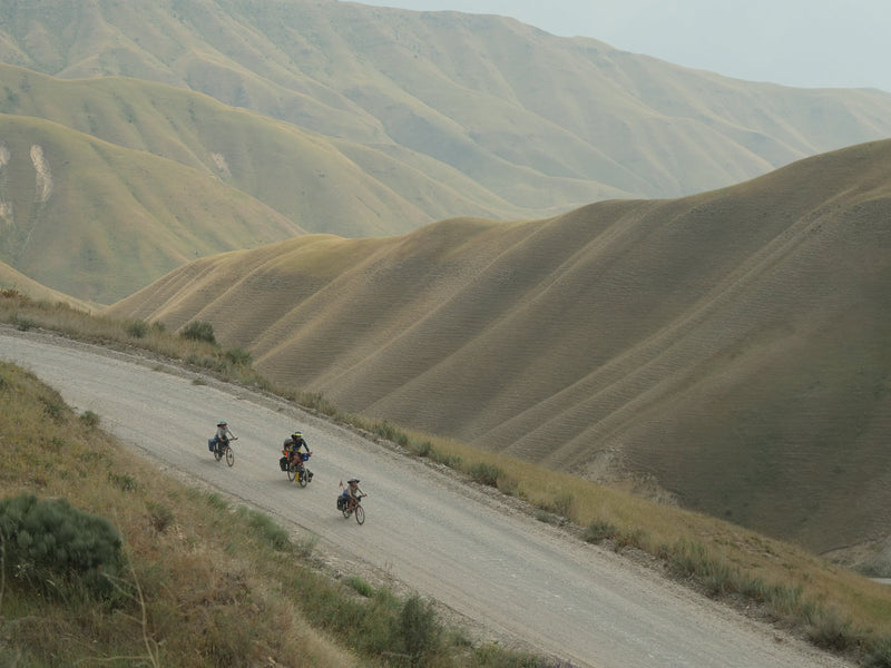 Cyclists riding up a winding gravel road through steep, rolling mountain hills.