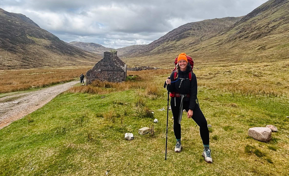 A smiling hiker in an orange beanie, black gear, and a red‑strapped backpack pauses with a trekking pole on a grassy trail dotted with rocks. An old stone cottage sits beside the dirt path, framed by a wide, rugged mountain valley under cloudy skies.