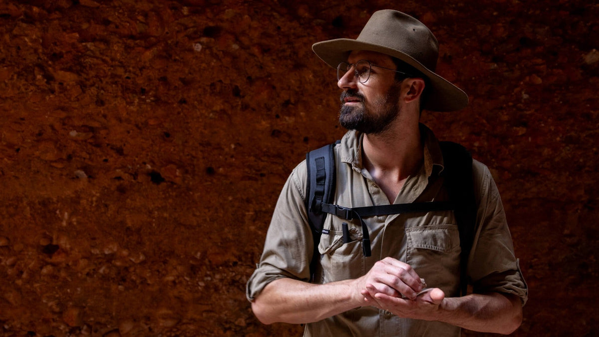 A man wearing a hat, glasses, and a backpack stands in front of a reddish-brown rock wall, looking to the right.