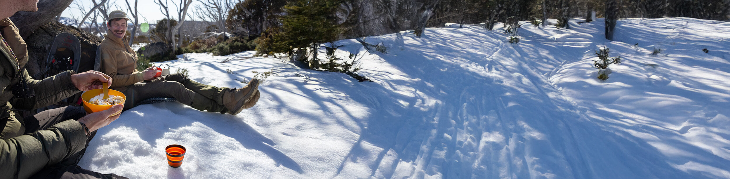 Two friends eating outdoors in the snow while laughing together.