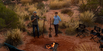 A Woman and a man standing by a campfire in the Australian desert at dawn.