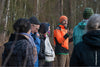 A group of volunteers listening to a man in an orange jacket giving instructions during an outdoor forest activity.