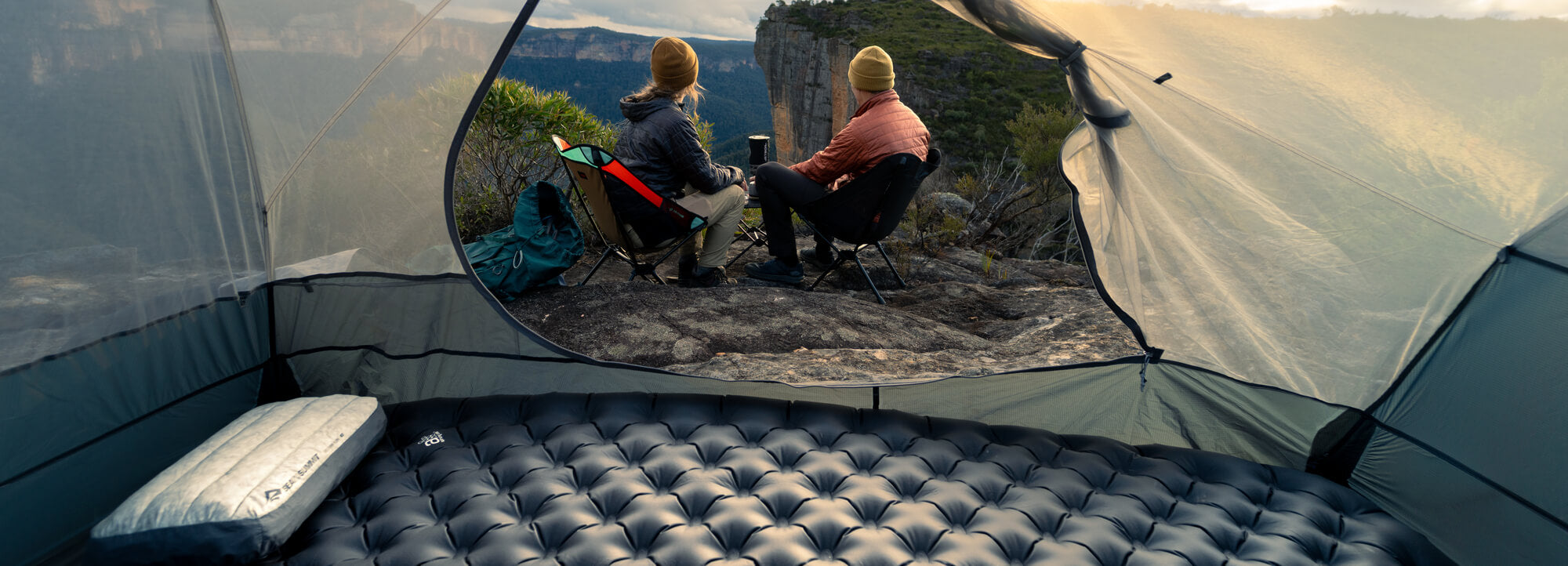 Two people sitting outside a tent in a colder season. Inside a tent a black sleeping mat - the well insulated Ether Light XR Pro mat - is visible.