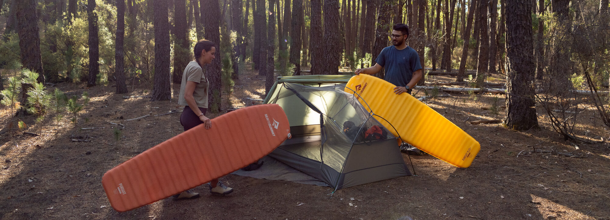 A woman and a man are holding the Pursuit and Pursuit Plus sleeping mats. They are about to put them into the Telos TR2 tent in a forest.